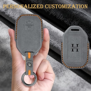 May include: Gray key fob cover with orange stitching and accents, featuring a key ring. The cover has a leather-like texture and the text "PERSONALIZED CUSTOMIZATION" is at the top. A second cover is shown with the initials "II HRNRY".