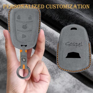 Puede incluir: Funda para llave de coche de ante gris con costuras naranjas y una correa naranja a juego. La funda tiene símbolos de llave en relieve y la palabra "Gospel" en la parte posterior. El texto "PERSONALIZACIÓN PERSONALIZADA" está en la parte superior.