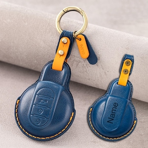 Könnte beinhalten: Zwei blaue Autoschlüsselhüllen aus Leder mit gelben Nähten und gelben Lederakzenten. Eine Hülle zeigt geprägte Tasten-Symbole, während die andere das Wort Name auf der Vorderseite eingeprägt hat. Beide haben einen messingfarbenen Schlüsselring.