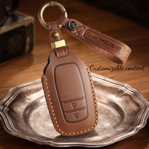 May include: A brown leather key fob with a key ring and a strap engraved with "David 1995.06.01". The key fob has two buttons and is stitched with a contrasting thread. The words "Customizable content" are also visible.