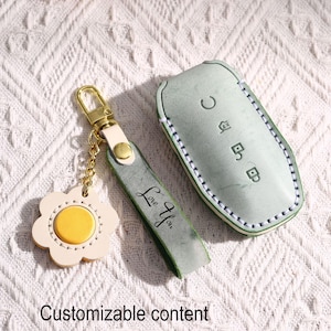 May include: A light green leather car key fob cover and matching keychain set. The key fob cover has white stitching and embossed symbols. The keychain features a flower-shaped charm with a yellow centre and the words "Love You" on the strap.