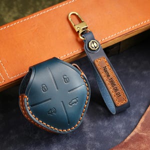 May include: A teal leather car key fob cover and matching keychain. The key fob cover has embossed icons and brown stitching. The keychain has a leather strap with the text "Name.1995.06.01" and a gold-colored clasp.