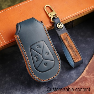 May include: A blue leather car key fob cover with orange stitching and a matching blue leather keychain strap. The strap features a brown leather inlay with the engraved text Name.1995.06.01 and a gold-toned metal lobster clasp hardware.