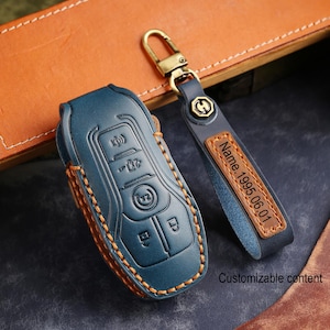 Peut inclure: Un étui de clé de voiture en cuir bleu et un porte-clés assorti avec des éléments dorés. L'étui de clé a des boutons en relief et des coutures orange. Le porte-clés porte l'inscription "Name.1995.06.01".