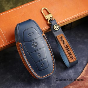 Puede incluir: Una funda para llave de coche de cuero azul marino con un llavero a juego. La funda tiene un borde cosido marrón y un diseño en relieve. El llavero tiene un cierre dorado y una etiqueta de cuero con el texto "Name 1995.06.01".