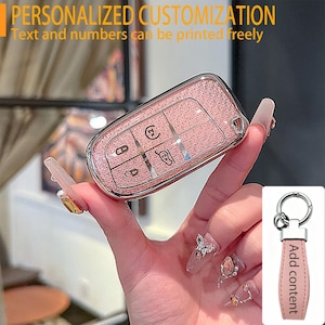 May include: A rose gold glitter car key fob cover with a glossy finish and silver-toned button icons. The cover features a rectangular shape with rounded edges. A separate light pink leather keychain strap with a silver ring is also shown.
