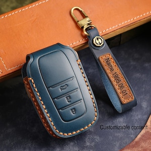 Puede incluir: Una funda de cuero azul oscuro para llave de coche con costuras naranjas y una correa de llavero a juego. La correa tiene una inserción de cuero tostado con el texto grabado Name. 1995.06.01 y un cierre de mosquetón de metal latonado.