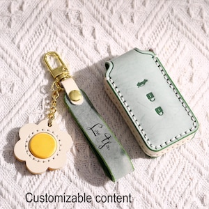 May include: A light green leather key fob cover and matching keychain set. The key fob cover has white stitching and a car symbol. The keychain features a daisy-shaped charm with a yellow center and the text "Love You."