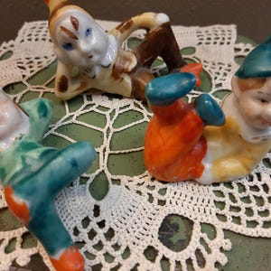 May include: Four small, colorful ceramic gnome figurines. One gnome is lying down with brown and tan clothing. Another is in a green outfit, and a third is wearing orange pants. The fourth gnome has a blue hat and is wearing yellow.