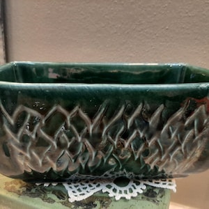 Puede incluir: Una jardinera rectangular de cerámica verde oscuro con un patrón texturizado similar a un tejido en los lados. La jardinera se asienta sobre un tapete de encaje blanco decorativo. La jardinera tiene un acabado brillante.