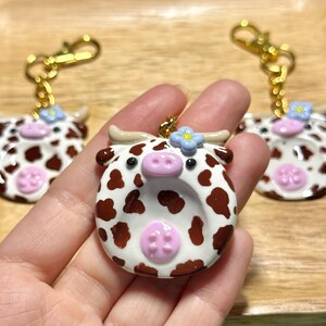 May include: Handmade cow-shaped keychains with white, brown, and pink details. Each keychain features a gold-colored clasp and a small blue flower accent. The cow keychains are held in a hand.