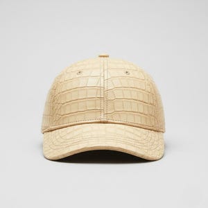 Könnte beinhalten: Beige Baseball Cap mit einem strukturierten, krokodilartigen Muster. Die Kappe hat einen gebogenen Schirm und einen Knopf oben. Der Hut ist eine neutrale Farbe und scheint aus Leder oder einem ähnlichen Material zu sein.