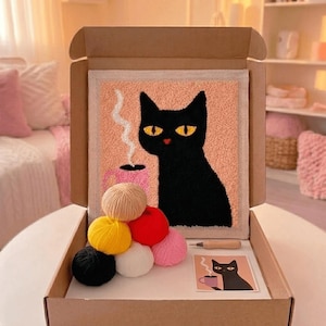 Op de afbeelding: Een punch needle set in een kartonnen doos met een vierkant stoffen paneel met een zwarte kat en een roze mok met stoom. Inbegrepen zijn zeven bollen garen in zwart, geel, beige, rood, wit en roze, een houten punch needle en een referentiekaart.