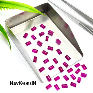 Puede incluir: Una colección de pequeñas piedras preciosas rectangulares de color magenta vibrante esparcidas en una bandeja de clasificación de metal plateado. Un par de pinzas de metal plateado descansa cerca. El texto NaviGemsIN está impreso en negro abajo.
