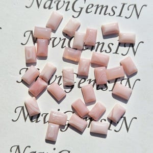Puede incluir: Una colección de piedras preciosas rectangulares y pulidas en varios tonos de rosa. Las piedras están dispersas sobre una superficie blanca con texto en cursiva en el fondo. Las piedras tienen un aspecto liso y brillante.