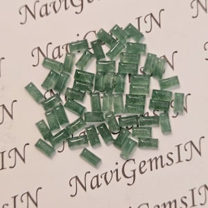 Könnte beinhalten: Eine Ansammlung kleiner, rechteckiger, durchscheinender grüner Edelsteine, die auf einer weißen Oberfläche verstreut sind. Die Edelsteine haben unterschiedliche Größen und Formen und sind poliert. Der Text "NaviGemsIN" ist im Hintergrund sichtbar.