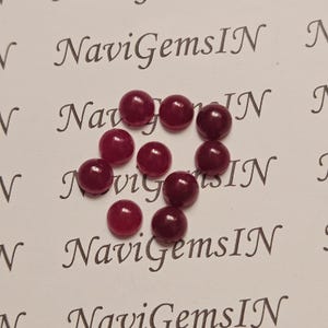 Könnte beinhalten: Zehn runde, polierte, tiefrote Edelsteine liegen auf einer weißen Oberfläche. Der Text "NaviGemsIN" ist in kursiver Schrift mehrfach abgedruckt. Die Edelsteine haben eine glatte, reflektierende Oberfläche.