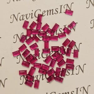 Puede incluir: Una colección de pequeñas gemas rectangulares de color rubí dispersas sobre una superficie blanca. Las gemas son translúcidas y tienen un aspecto pulido. La imagen incluye el texto "NaviGemsIN" en cursiva.