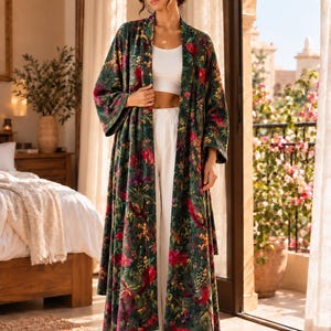 May include: A long, open-front robe in a dark green velvet fabric featuring a vibrant, multi-colored floral and botanical print. The robe has wide, loose sleeves and a relaxed, floor-length silhouette, worn over a white crop top and pants.