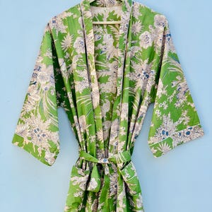 May include: A green floral print robe with a matching belt. The robe features a white trim and a large floral pattern in white and blue. The robe is hanging on a wooden hanger against a light blue background.