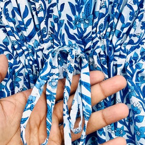 May include: Close-up of a white and blue floral patterned garment. The fabric features a repeating design of stylized flowers and leaves in varying shades of blue. A drawstring is visible, held by a hand, adding a functional and decorative element.