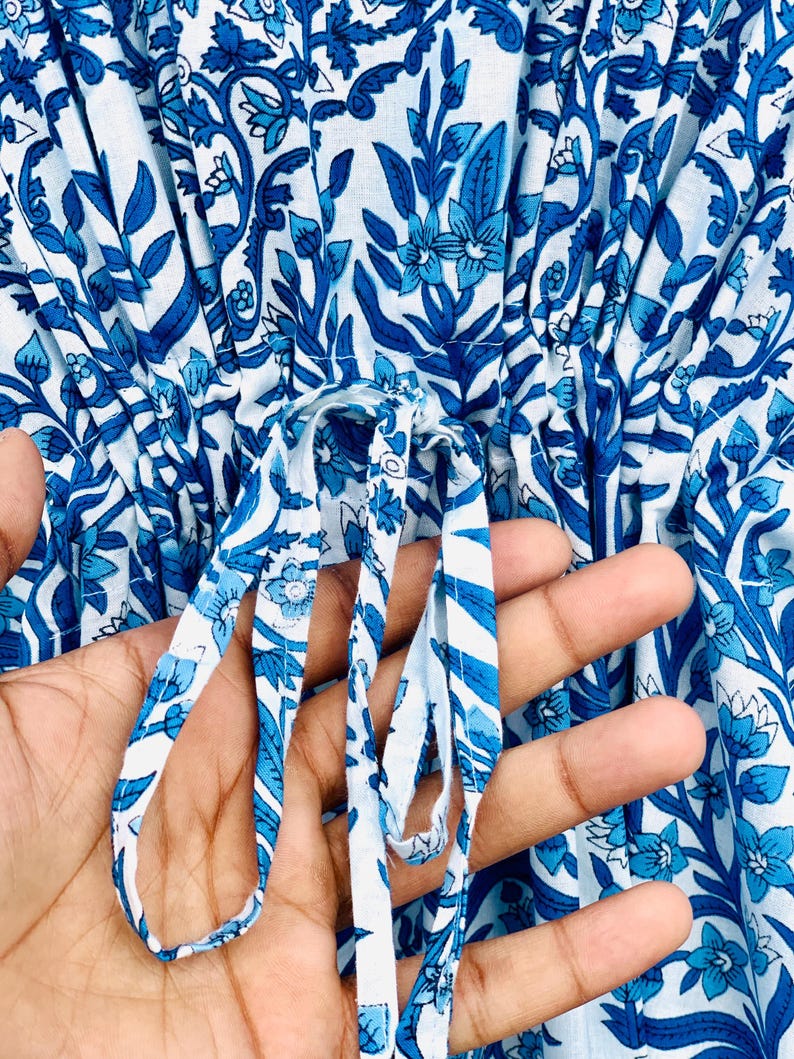 May include: Close-up of a white and blue floral patterned garment. The fabric features a repeating design of stylized flowers and leaves in varying shades of blue. A drawstring is visible, held by a hand, adding a functional and decorative element.