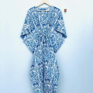 May include: A blue and white patterned kaftan dress hanging on a wooden hanger. The dress features a floral design in shades of blue against a white background. The kaftan has wide sleeves and a tie at the waist. The garment is made of a lightweight fabric.