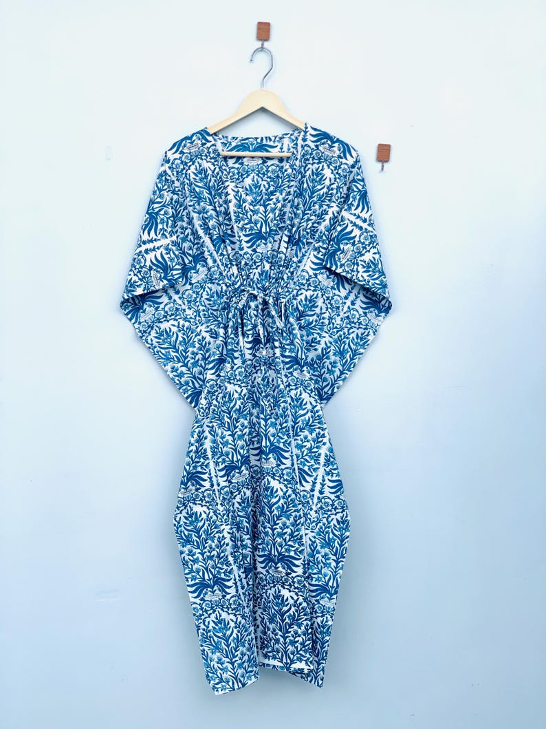May include: A blue and white patterned kaftan dress hanging on a wooden hanger. The dress features a floral design in shades of blue against a white background. The kaftan has wide sleeves and a tie at the waist. The garment is made of a lightweight fabric.