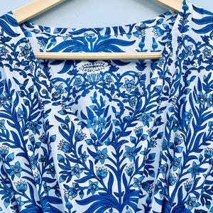 May include: White and blue floral patterned fabric, likely a garment, hangs on a wooden hanger. The design features stylized blue flowers and foliage against a white background. The fabric is visible from the neckline down.