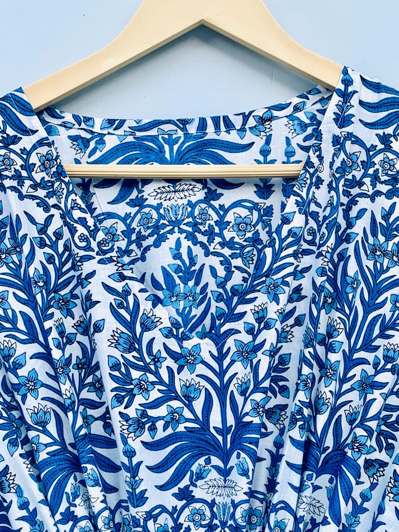 May include: White and blue floral patterned fabric, likely a garment, hangs on a wooden hanger. The design features stylized blue flowers and foliage against a white background. The fabric is visible from the neckline down.