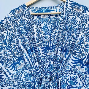 May include: A blue and white patterned caftan dress with a floral design, hanging on a wooden hanger. The dress features a V-neck and a drawstring waist. The fabric is a light, flowing material, perfect for warm weather.