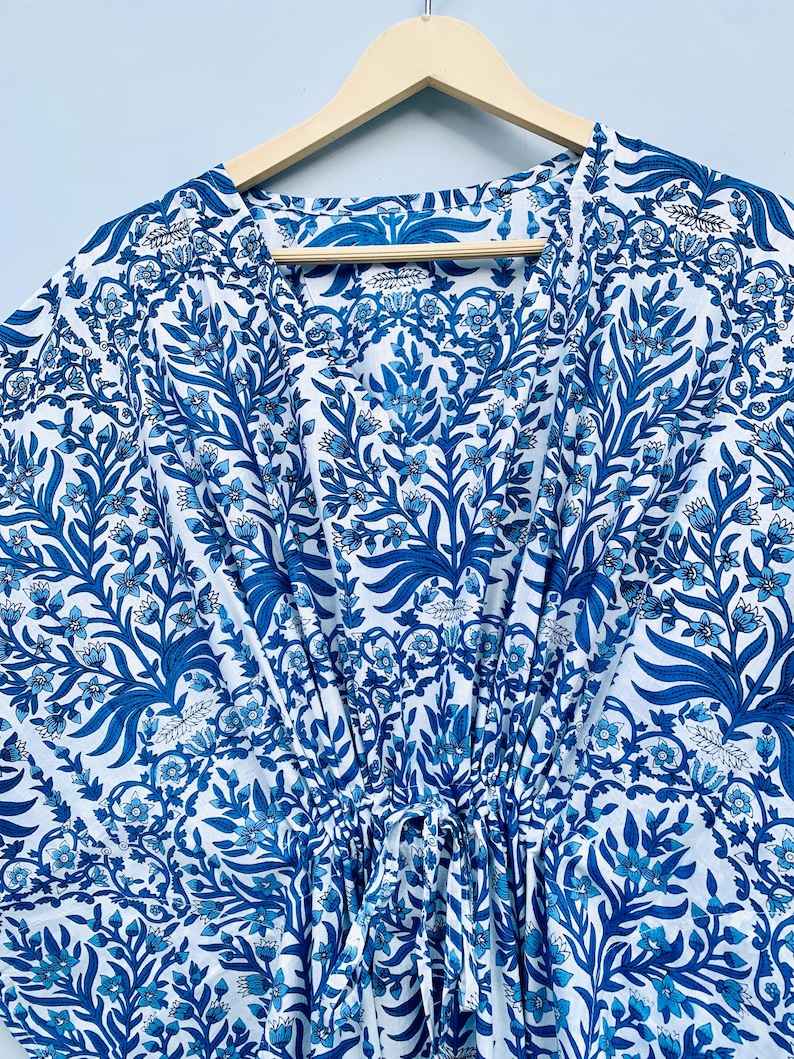 May include: A blue and white patterned caftan dress with a floral design, hanging on a wooden hanger. The dress features a V-neck and a drawstring waist. The fabric is a light, flowing material, perfect for warm weather.