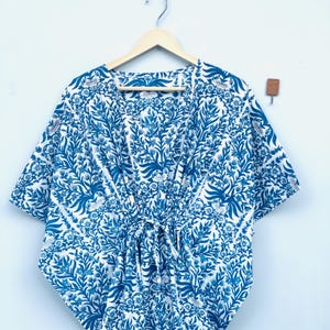 May include: A blue and white patterned kaftan dress with a drawstring waist hangs on a wooden hanger. The fabric features a floral design in shades of blue against a white background. The dress has short sleeves and a V-neck.