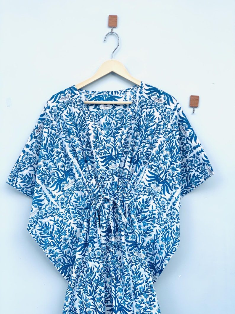 May include: A blue and white patterned kaftan dress with a drawstring waist hangs on a wooden hanger. The fabric features a floral design in shades of blue against a white background. The dress has short sleeves and a V-neck.