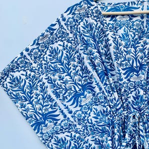 May include: A blue and white patterned kaftan dress with a floral design. The dress is hanging on a wooden hanger. The fabric features a repeating pattern of stylized leaves and flowers in shades of blue against a white background.