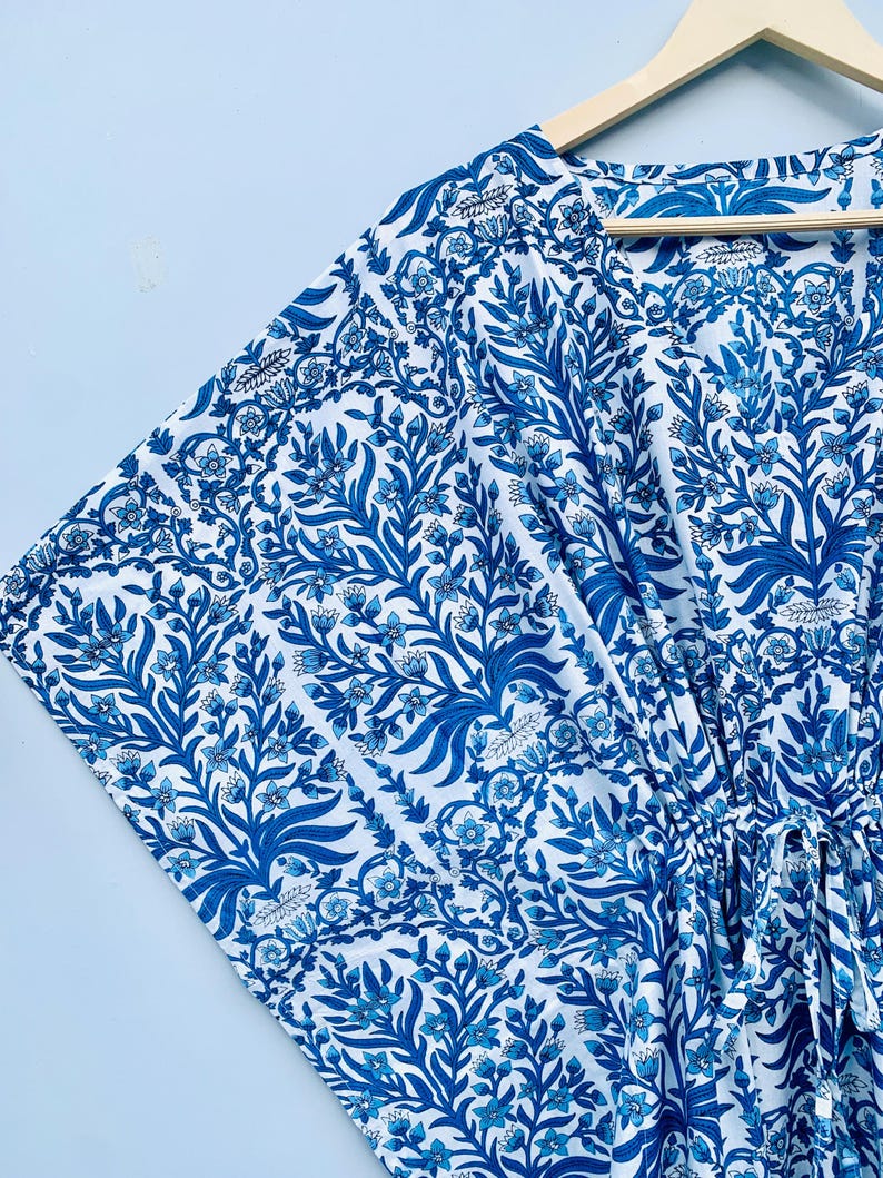 May include: A blue and white patterned kaftan dress with a floral design. The dress is hanging on a wooden hanger. The fabric features a repeating pattern of stylized leaves and flowers in shades of blue against a white background.