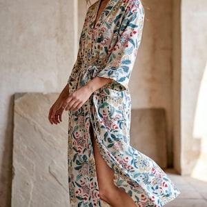 Könnte beinhalten: Langer Morgenmantel mit Blumenmuster in Creme, Rot, Blau und Grün. Der Morgenmantel hat einen Bindegürtel, Dreiviertelärmel und weiße Paspeln. Der Morgenmantel ist knielang und hat einen hohen Schlitz an der Seite.