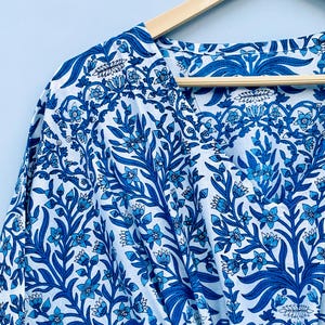 May include: A white and blue floral patterned garment, possibly a robe or kimono, hangs on a wooden hanger. The fabric features a detailed design of blue flowers and leaves against a white background. The V-neckline is visible.
