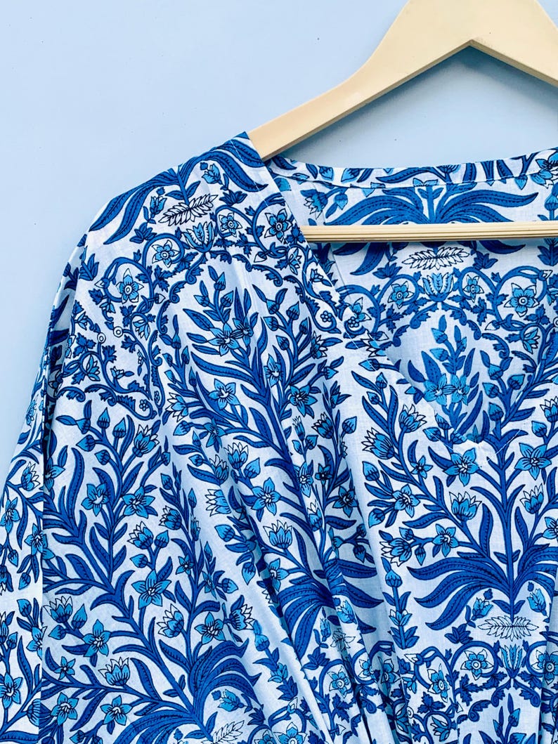 May include: A white and blue floral patterned garment, possibly a robe or kimono, hangs on a wooden hanger. The fabric features a detailed design of blue flowers and leaves against a white background. The V-neckline is visible.