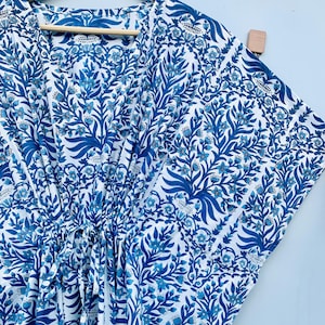 May include: A white and blue patterned caftan dress with a V-neckline, hanging on a wooden hanger. The fabric features a repeating floral design in shades of blue. The dress has a drawstring at the waist.