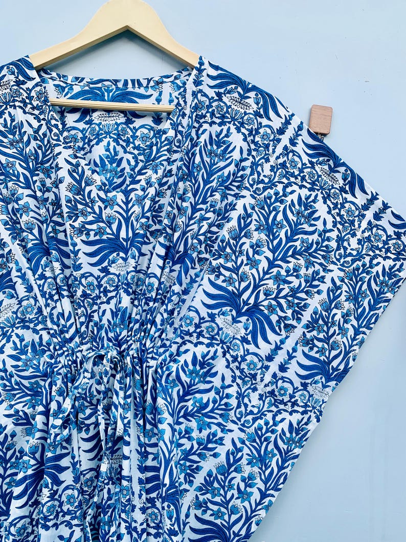 May include: A white and blue patterned caftan dress with a V-neckline, hanging on a wooden hanger. The fabric features a repeating floral design in shades of blue. The dress has a drawstring at the waist.