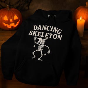 Peut inclure: Un sweat à capuche noir avec un motif blanc d'un squelette dansant portant un chapeau de sorcière. Le texte "DANCING SKELETON" est au-dessus. Deux citrouilles sculptées et une bougie allumée sont en arrière-plan.