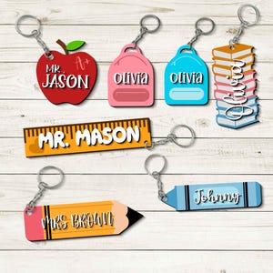 May include: A collection of personalized keychains in various shapes, including an apple, backpacks, books, a ruler, a pencil, and a crayon. Each keychain features a name, such as "Jason," "Olivia," "Mr. Mason," "Mrs. Brown," and "Johnny."