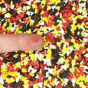May include: A close-up of a finger holding a small red Mickey Mouse-shaped sprinkle. The background is filled with a colorful assortment of similar sprinkles in red, yellow, black, and white. These sprinkles are likely for decorating baked goods.