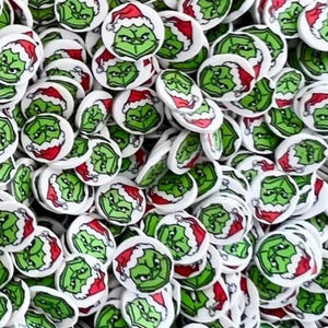 Pode incluir: Uma foto em close-up de uma pilha de pequenos enfeites de Natal redondos. Cada disco branco apresenta um rosto verde do Grinch usando um chapéu de Papai Noel vermelho e branco. Os enfeites estão espalhados, criando um design festivo de feriado.