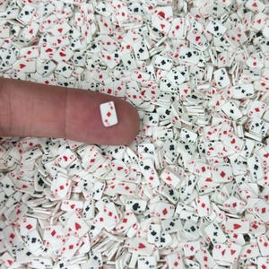 May include: Miniature playing card charms in a variety of suits, including hearts, diamonds, clubs, and spades. The cards are white with red and black suit symbols. One diamond card is held by a finger.