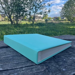 Puede incluir: Un libro rectangular con una cubierta de color turquesa brillante, lisa y sólida. El libro está cerrado, mostrando la gruesa pila de páginas de papel blanco a lo largo del borde lateral. La cubierta tiene un acabado mate y color uniforme.