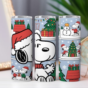 Puede incluir: Tres vasos térmicos blancos con Snoopy con gorro de Papá Noel, decorados con paneles de cómic navideños. Los paneles muestran a Snoopy con un muñeco de nieve, un árbol de Navidad y regalos, sobre un fondo azul claro con copos de nieve.