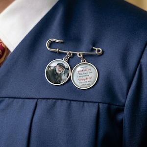 May include: A silver-colored pin with two circular charms. One charm features a photo of a man, and the other has text that reads, "On your graduation day, know that I am walking beside you every step of the way."