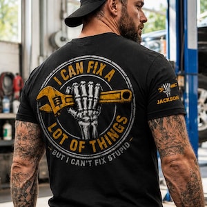 May include: A black short-sleeve t-shirt featuring a graphic on the back of a skeleton hand holding a yellow wrench inside a circular border. The text reads I CAN FIX A LOT OF THINGS BUT I CAN'T FIX STUPID. A smaller wrench logo with the text JACKSON is on the sleeve.
